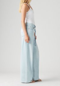 Vista lateral de una persona que lleva unos pantalones vaqueros de pierna ancha en azul claro con un cinturón y una camiseta sin mangas blanca, de pie descalza sobre un fondo liso.