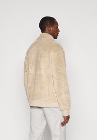 Hustá béžová bunda s vysokým golierom, mäkkou textúrou a elastickými manžetami. Nosí sa s šedými teplákmi, zobrazené zozadu.