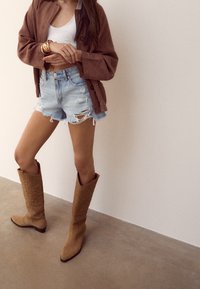 Femme portant une veste marron, un débardeur blanc, un short en jean déchiré, des bracelets dorés et des bottes de cowboy marron montantes, debout à l'intérieur.