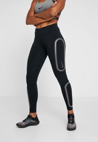 Žena v černých legínách Nike s šedými reflexními paneli a šedým sportovním podprsenkou Nike, stojící se složenýma rukama na bílém pozadí.