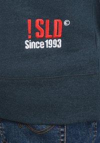 Donkerblauwe trui met geribbelde zoom, met geborduurde tekst "! SLD ©" in het rood en "Since 1993" in het wit. Denim jeans zijn zichtbaar eronder.