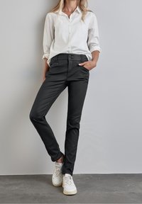 Musta räätälöity housut sujuvalla pinnalla, joissa on kaksi etutaskua, vetoketju- ja nappisulku, päällä valkoinen kauluspaita ja lenkkarit.