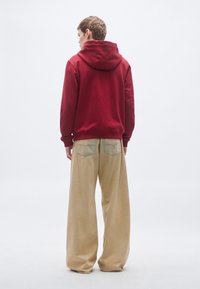 Bordová mikina s kapucňou so sťahovacou šnúrkou, v kombinácii s voľnými béžovými nohavicami so širokými nohavicami, ktoré majú zadné vrecká a jemné šijacie detaily.