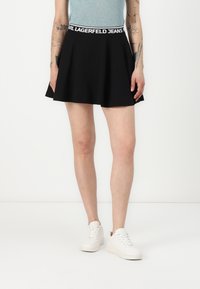 Svart skater skjørt med bred elastisk midjebånd med en hvit logo, laget av glatt stoff, som rekker til midt på låret, stylet med hvite joggesko.