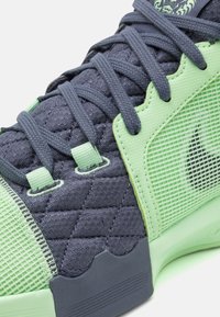 Gros plan sur une basket verte et gris foncé montrant un tissu matelassé, un panneau en maille et des lacets tissés noués sur une languette rembourrée avec un patch logo.