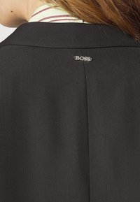 Musta räätälöity takki, jossa on teksturoitu kangas ja selässä kaulan tienoilla näkyvä "BOSS"-logo.