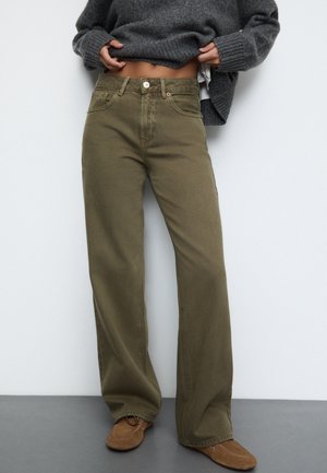 Personne portant un jean large vert olive, des chaussures marron décontractées et un pull gris foncé relevé pour dévoiler le ventre.