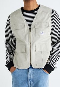 Gilet beige con zip anteriore e quattro tasche frontali, indossato sopra una camicia a righe bianca e nera a maniche lunghe, abbinato a jeans azzurri chiari.