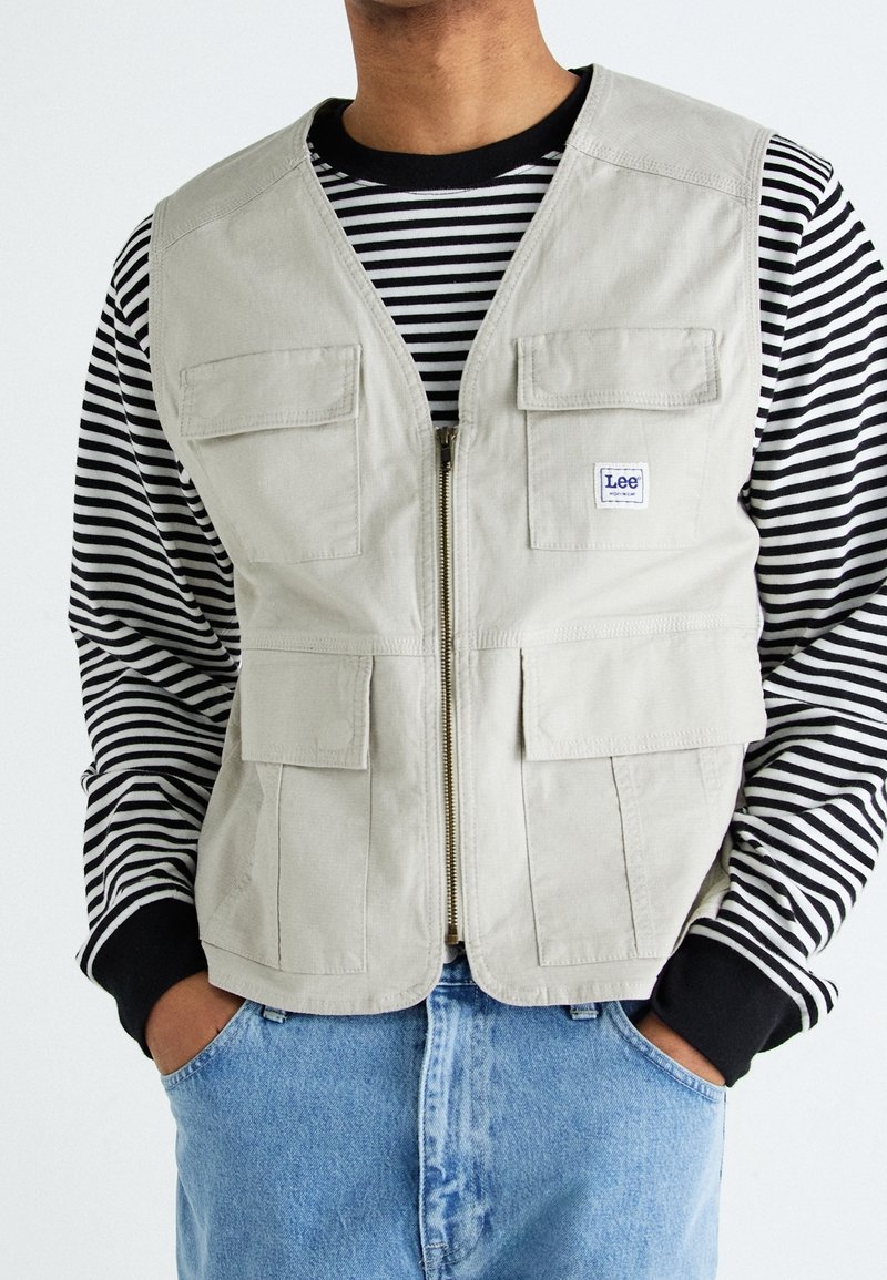 Gilet beige con zip anteriore e quattro tasche frontali, indossato sopra una camicia a righe bianca e nera a maniche lunghe, abbinato a jeans azzurri chiari.