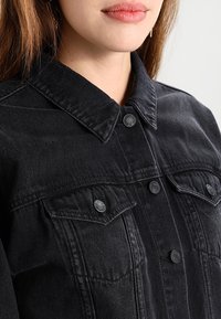 Svart denimjacka med klassisk krage, framknäppning med knappar och två bröstfickor med svarta knappdetaljer. Tyget är strukturerat.