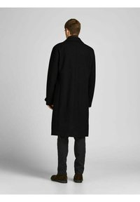 Svart ullrock med lång design, med rak passform och breda slag, bärs över mörka byxor och bruna mockaskor.