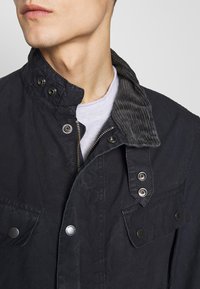 Black jacket featuring a corduroy collar, snap buttons, and zipper. Made from textured fabric with two front pockets.