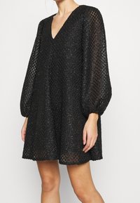 Mujer vestida con un vestido negro de escote en V, con mangas largas y transparentes abullonadas y tela con patrón de diamante texturizado.