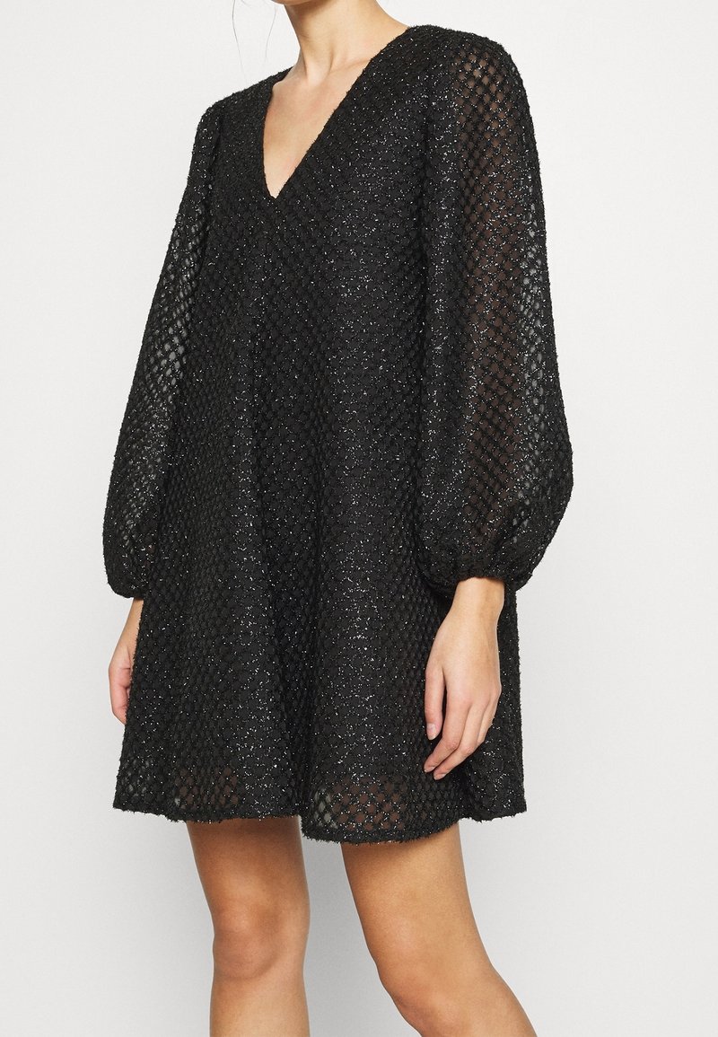 Mujer vestida con un vestido negro de escote en V, con mangas largas y transparentes abullonadas y tela con patrón de diamante texturizado.