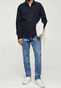 Chemise bleu marine à manches longues avec boutons, jean bleu clair et baskets blanches. Un pull crème drap é sur le bras. Matière en coton, coupe décontractée.