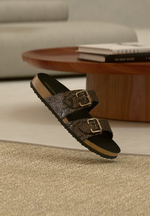 Brun tekstureret sandal med dobbelt rem og gyldne spænder på tæppebelagt gulv, træbord og bøger i baggrunden.