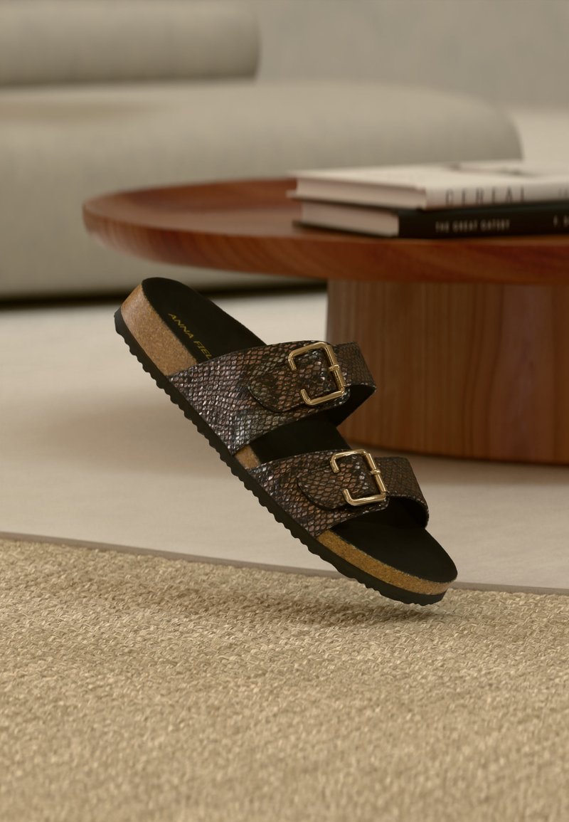 Sandale marron texturée à double bride avec boucles dorées sur sol moquetté, table en bois et livres en arrière-plan.