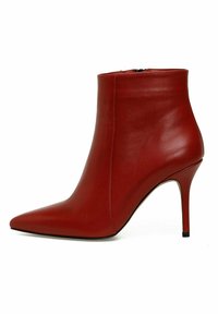 Bottine en cuir rouge avec un bout pointu, un talon aiguille et une texture lisse. Dispose d'une fermeture éclair latérale pour faciliter l'enfilage.