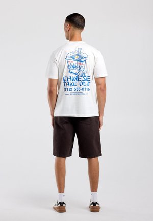 Man draagt een wit T-shirt met blauwe en rode afbeelding van een Chinese afhaaldoos en tekst, gecombineerd met zwarte korte broek, witte sokken en sneakers.