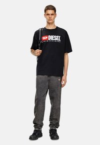 Melnas kokvilnas T-krekls ar baltu un sarkanu "DIESEL" zīmolu, apvienots ar tumši pelēkiem džinsiem ar elastīgiem uzmaviem un sānu logotipu. Melni apavi.