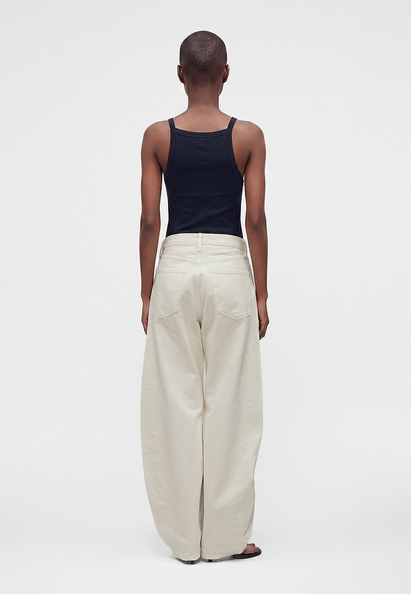 Personne portant un haut sans manches noir et un pantalon large écru, debout de dos devant un fond gris uni.