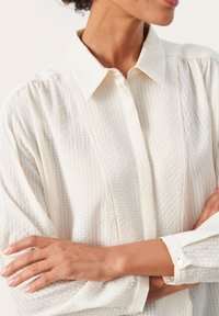 Bílá texturovaná blůza s límečkem, zapínáním na knoflíky vpředu a dlouhými rukávy, která má jemné vyvýšené vzory a knoflíky na manžetách.