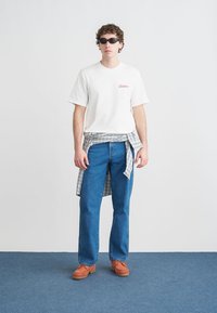 Λευκό T-shirt με κοντά μανίκια και λογότυπο "Dickies", μπλε τζιν, καρό πουκάμισο δεμένο στη μέση, γυαλιά ηλίου και καφέ παπούτσια με ανάγλυφες σόλες.