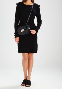 Svart långärmad klänning med volangdetaljer, i kombination med en svart axelremsväska med guldspänne och svarta sandaler.