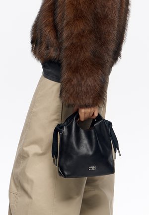 Osoba ubrana w brązową futrzaną kurtkę i beżowe spodnie trzyma małą czarną skórzaną torebkę z logo "Bimba Y Lola".