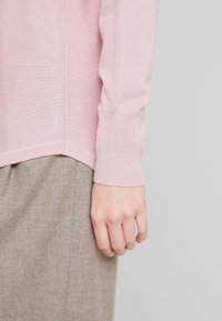 Maglione rosa chiaro lavorato a maglia con orlo arrotondato e polsini a coste, abbinato a pantaloni marroni con texture. Mano visibile appoggiata sui pantaloni.