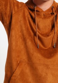 Sudadera marrón hecha de una tela suave y texturizada con un bolsillo en la parte frontal. Presenta una capucha con cordón y acentos metálicos en los cordones.