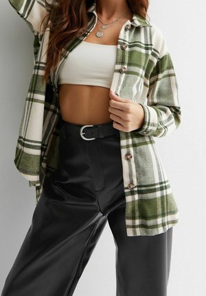 Vrouw draagt een groen-wit geruite blouse over een wit cropped topje, zwarte hoge taille broek met een zilverkleurige gesp en een ketting.