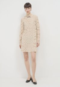 Spitzenkleid in hellbeige mit langen Ärmeln, floralem Muster und einem schwarzen Schleifenakzent am Ausschnitt. Das Model trägt schwarze High Heels.