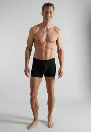 Lächelnder, oberkörperfreier Mann mit athletischer Figur, barfuß stehend und schwarze Boxershorts tragend vor hellem, einfachem Hintergrund.