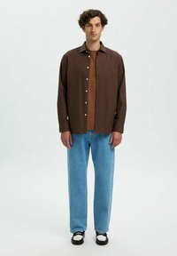 Braunes Hemd mit Knöpfen über einem braunen Top, kombiniert mit hellblauen Jeans und schwarz-weißen Schuhen. Das Hemd hat eine glatte Textur.