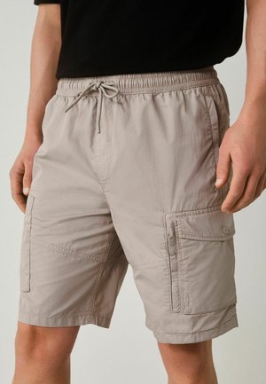 Man wearing beige cargo shorts with multiple pockets and a drawstring waist, paired with a black shirt, shown from waist to knees.