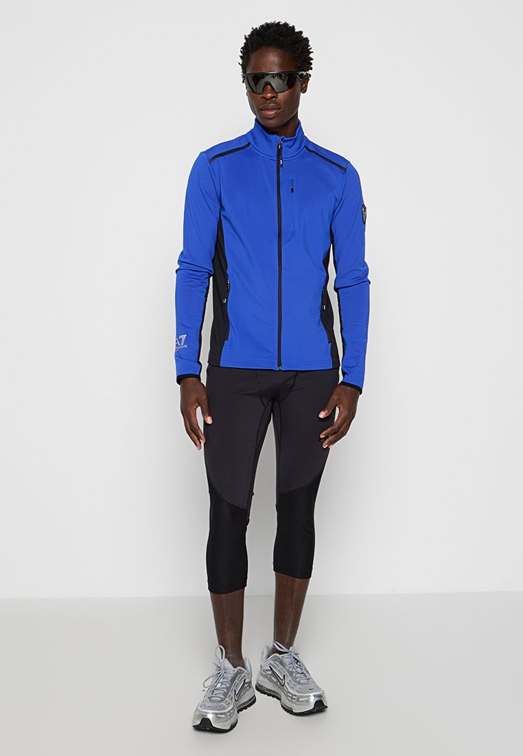 Giacca sportiva blu con pannelli laterali neri, tasche con zip e colletto alto, abbinata a leggins neri aderenti e scarpe da corsa argentate.