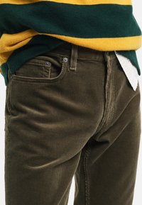 Brązowe spodnie w koszykowej strukturze z teksturowanym wykończeniem, z guzikowym rozporkiem i przednimi kieszeniami, zestawione z swetrem w tęczowy pasek.