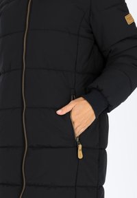 Giubbotto a sacco nero con cuciture orizzontali, zip frontale e tasca laterale. I polsini sono a coste; i dettagli includono un'etichetta in pelle beige.