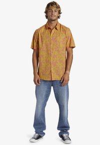 Korte mouwen, overhemd met knoopsluiting in het geel met een roze abstract patroon; gecombineerd met lichtblauwe jeans en zwarte sneakers.