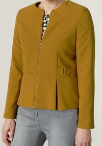 Femme portant une veste ajustée jaune moutarde avec une fermeture éclair et un détail boucle, associée à un jean gris clair et un top imprimé animalier en dessous.