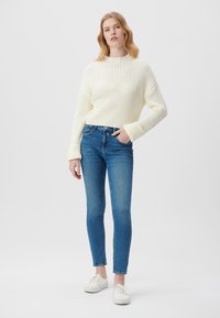 Cremfarbener, gerippter Strickpullover mit hohem Kragen, kombiniert mit blauen Skinny-Jeans. Das Model trägt weiße Sneakers und hat die Hände in den Taschen. Schlichter Hintergrund.