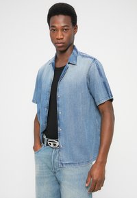 Πουκάμισο από τζιν με κοντά μανίκια σε ανοιχτό μπλε, με κουμπώματα μπροστά, γιακά με αιχμηρές άκρες και άνετη εφαρμογή. Συνδυάζεται με σκούρα τζιν.