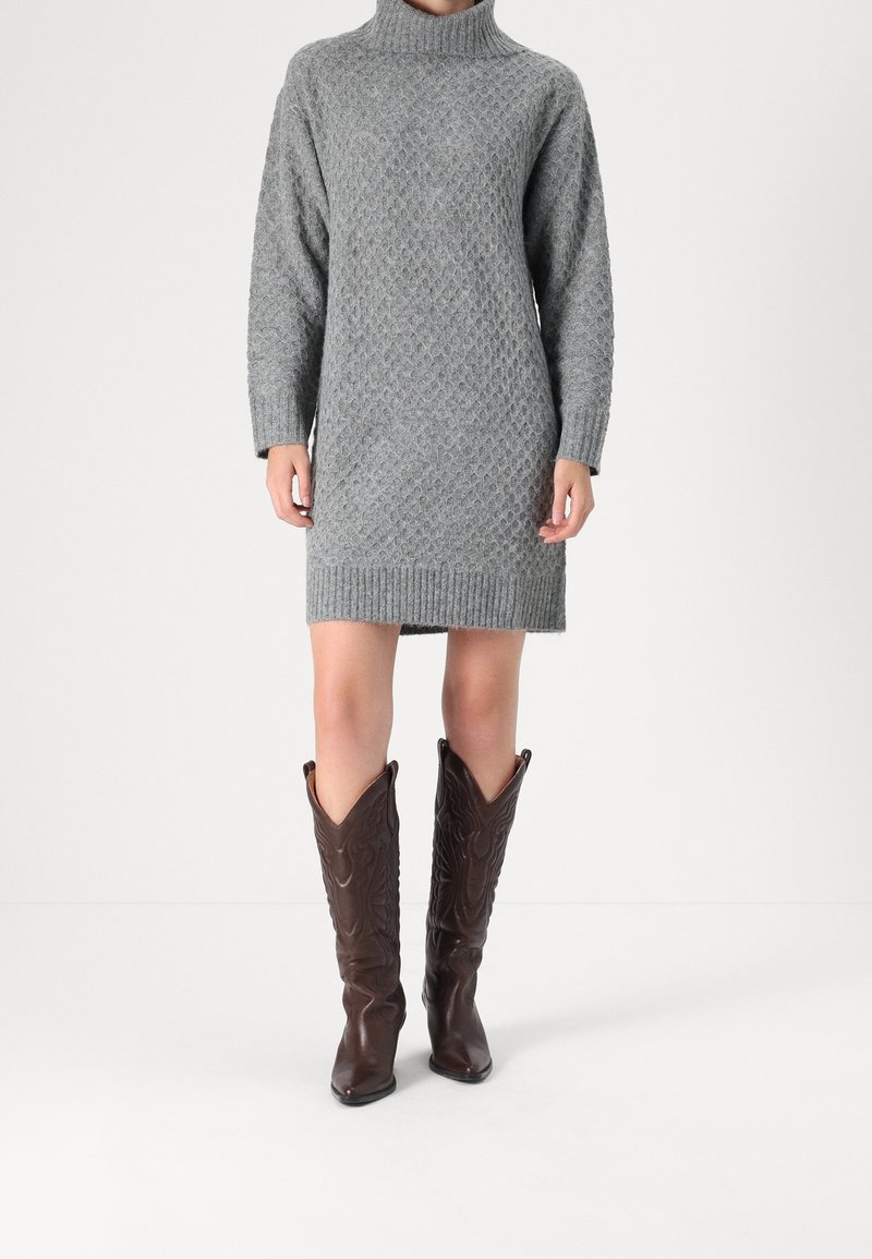 Šedé pletené šaty se stojáčkem, zdobené strukturovaným diamantovým vzorem a žebrovanými manžetami, spojené s hnědými kovbojskými botami.