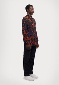 Maglietta nera a maniche lunghe con un motivo paisley colorato e dettagliato, abbinata a jeans scuri e sneaker bianche.