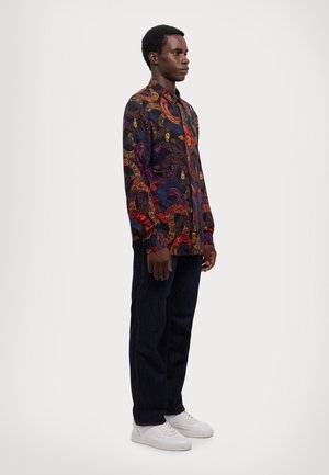 Maglietta nera a maniche lunghe con un motivo paisley colorato e dettagliato, abbinata a jeans scuri e sneaker bianche.