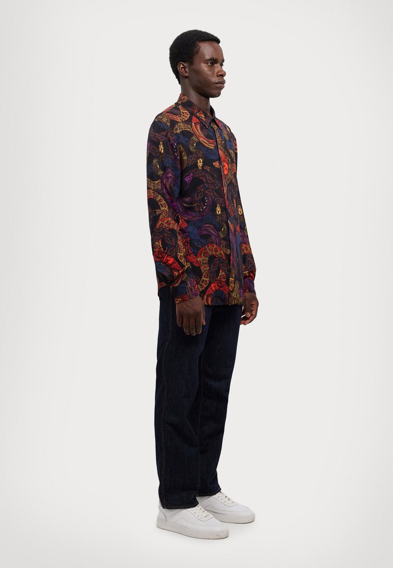 Maglietta nera a maniche lunghe con un motivo paisley colorato e dettagliato, abbinata a jeans scuri e sneaker bianche.