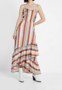 Sukienka maxi bez ramiączek z pionowymi, wielokolorowymi paskami, falbaną u dołu i zwiewnym krojem. Noszona z białymi botkami na obcasie.