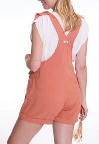 Rost-orange Latzhose aus einem leichten Stoff, mit verstellbaren, gebundenen Trägern, Seitentaschen und einem Etikett auf der Rückseite.