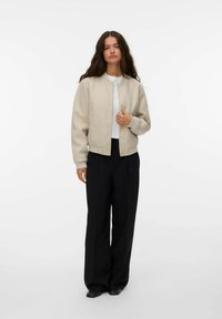Giacca bomber beige con polsini a costine, bottoni a pressione e una texture liscia, indossata sopra un top bianco e abbinata a pantaloni neri a gamba larga.